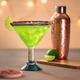 Margarita glass full of bright green liquid with a copper cocktail shaker and lime wedges behind it